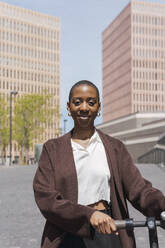 Smiling young businesswoman with electric push scooter on sunny day - PNAF05553