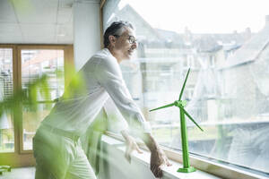 Nachdenklicher Geschäftsmann bei einem Windradmodell, das auf der Fensterbank eines Büros steht - UUF29143