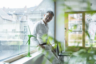 Lächelnder Geschäftsmann, der sich auf die Fensterbank lehnt und das Modell einer Windkraftanlage im Büro betrachtet - UUF29145