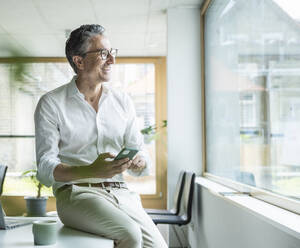 Happy businessman sitting on desk with smart phone looking out through window at workplace - UUF29156