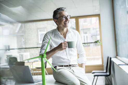 Thoughtful happy businessman holding coffee cup sitting on desk by wind turbine model in office - UUF29159