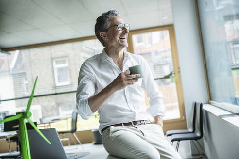 Älterer Geschäftsmann mit Kaffeetasse auf dem Schreibtisch sitzend und lachend im Büro - UUF29160
