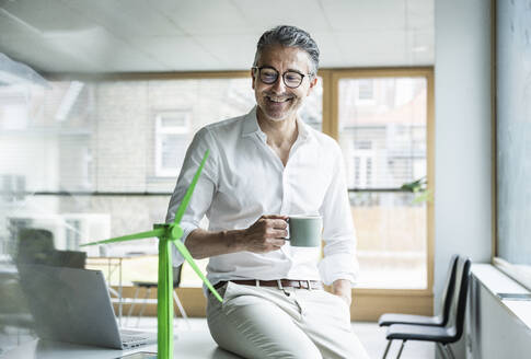 Smiling businessman holding coffee cup looking at wind turbine model in office - UUF29161