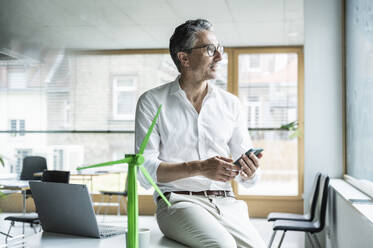 Thoughtful businessman sitting on desk with smart phone by wind turbine model at workplace - UUF29163