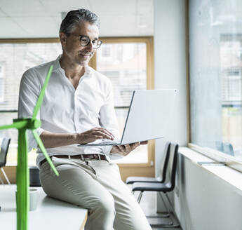 Businessman sitting on desk using laptop by wind turbine model in office - UUF29166