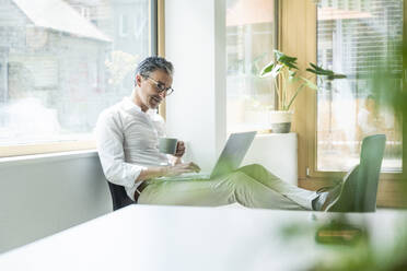 Confident businessman holding coffee cup using laptop working at office - UUF29171