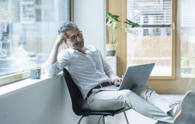 Confident businessman using laptop working near window at office - UUF29174