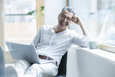 Happy businessman with laptop leaning head on window sill at office - UUF29179