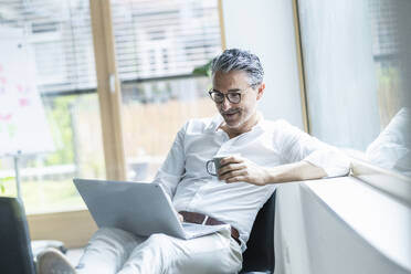 Mature businessman using laptop holding coffee cup leaning on window sill at office - UUF29182