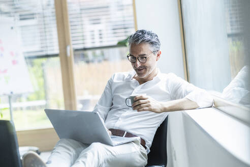 Älterer Geschäftsmann mit Laptop und Kaffeetasse auf der Fensterbank im Büro - UUF29182