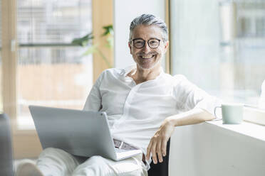 Happy businessman sitting with laptop leaning by window sill at office - UUF29186