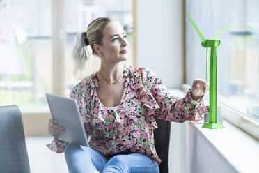 Businesswoman holding tablet PC looking at wind turbine model at office - UUF29193