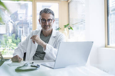 Selbstbewusster Arzt am Schreibtisch mit Laptop und Stethoskop in einer Klinik - UUF29228