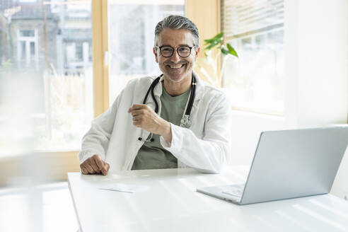 Lächelnder Arzt sitzt am Schreibtisch mit Laptop in einer Klinik - UUF29231