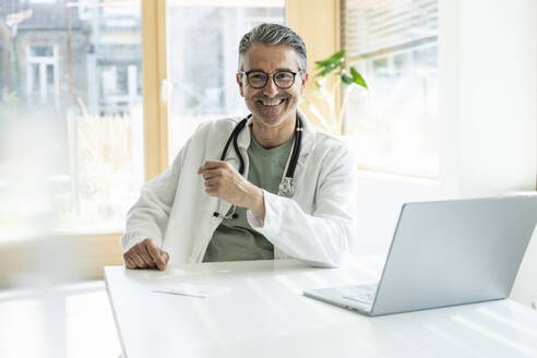 Smiling doctor sitting at desk with laptop in clinic - UUF29231