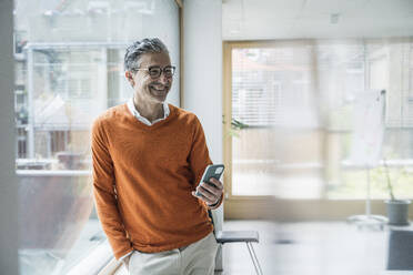 Smiling mature businessman holding smart phone leaning at glass window in office - UUF29237