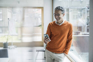 Smiling mature businessman using smart phone leaning at glass window in office - UUF29238