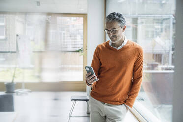 Mature businessman using smart phone leaning at glass window in office - UUF29239