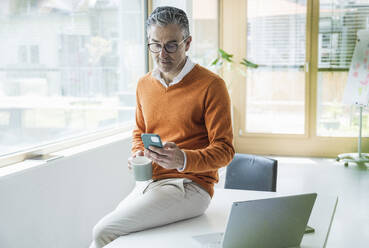 Businessman using smart phone sitting on table in office - UUF29245