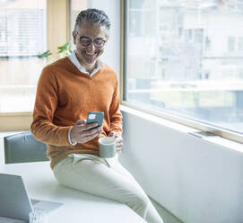 Smiling businessman using smart phone sitting on table in office - UUF29246