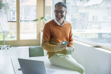 Smiling businessman holding smart phone and coffee cup sitting in office - UUF29247