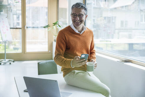 Lächelnder Geschäftsmann mit Smartphone und Kaffeetasse im Büro sitzend - UUF29247