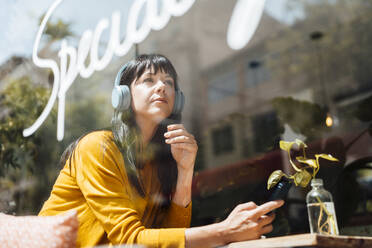 Contemplative woman wearing wireless headphones seen through glass - JOSEF19745