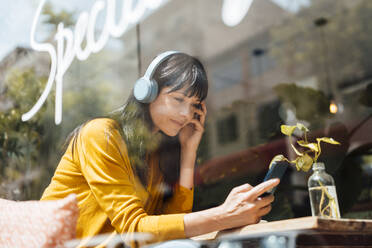 Mature woman wearing wireless headphones using smart phone in cafe - JOSEF19746