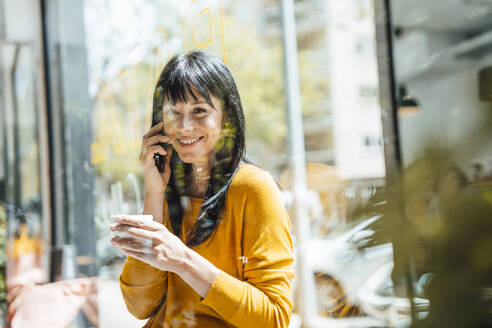 Happy mature woman with coffee cup talking on smart phone in cafe - JOSEF19753