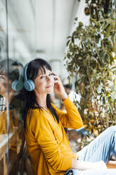 Smiling woman wearing wireless headphones listening to music by glass - JOSEF19772