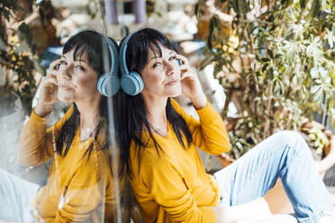 Smiling mature woman wearing wireless headphones listening to music by glass - JOSEF19773