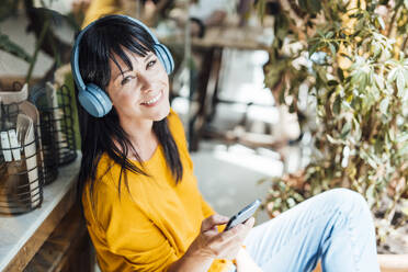 Glückliche reife Frau mit drahtlosen Kopfhörern, die mit einem Smartphone in einem Café sitzt - JOSEF19775