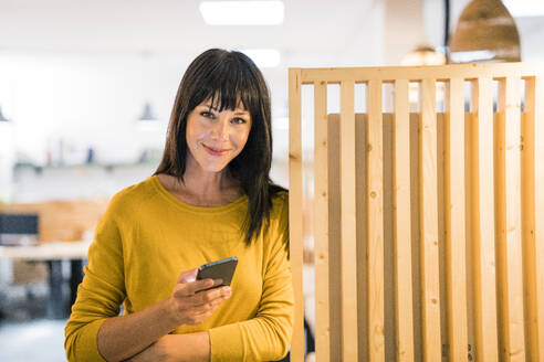 Lächelnde reife Geschäftsfrau im Büro stehend mit Smartphone - JOSEF19781