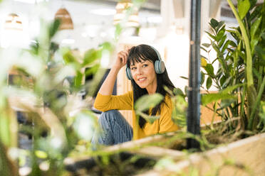 Thoughtful businesswoman wearing wireless headphones sitting in office - JOSEF19813