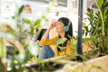 Happy businesswoman wearing wireless headphones sitting in office - JOSEF19814