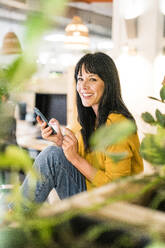 Smiling businesswoman sitting with credit card and mobile phone in office - JOSEF19818