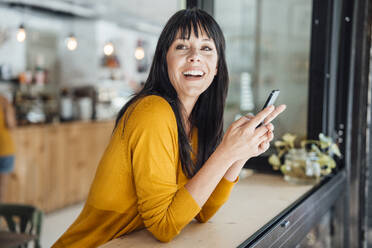 Glückliche Frau mit Smartphone, die sich in einem Café auf einen Tisch stützt - JOSEF19820