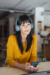 Smiling woman wearing wireless headphones standing with smart phone at table - JOSEF19824