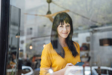 Smiling mature woman with smart phone seen through glass in cafe - JOSEF19834