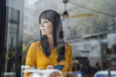 Thoughtful woman with smart phone seen through glass in cafe - JOSEF19835