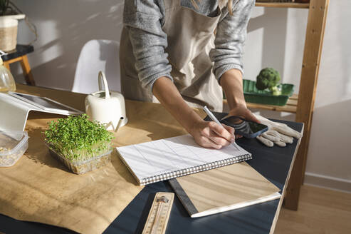 Woman writing in spiral book using smart phone at home - ALKF00358