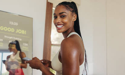 Happy and enthusiastic young woman using her smartphone and a smart mirror to enhance her fitness routine, utilizing advanced technology to achieve her fitness goals and maintain a healthy and active lifestyle. - JLPSF30501