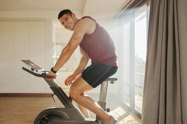 Young man enjoys smashing his goals in his indoor cycling workout. Athlete using his high-tech training equipment to get the most out of his home gym and push himself to new heights of fitness. - JLPSF30502