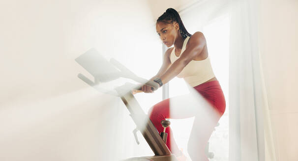 Focused young woman tackling her fitness training head-on, using a digital bike as part of her home workout routine to stay motivated and committed to achieving her fitness goals. - JLPSF30504