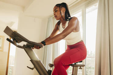 Determined young African woman actively pursuing her healthy lifestyle goals through digital fitness equipment, using her exercise bike to keep herself on track and focused on her routine. - JLPSF30508