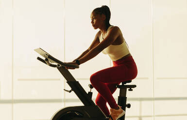 Sporty black female engaging in a healthy home workout routine, using a stationary bike as part of her fitness regimen. Young woman exercising with a focus on achieving her health and wellness goals. - JLPSF30515