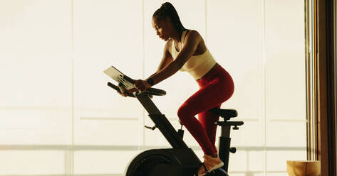 Fit African woman working out on a digital exercise bike, engaging in cardio to maintain her health and wellbeing. Woman using smart exercise equipment in her home fitness routine. - JLPSF30516