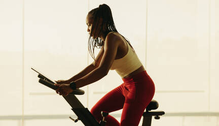 African American woman using an exercise bike to improve her cardio health and overall wellbeing. Young black woman engaging in fitness training in a gym, part of her wellness routine. - JLPSF30517