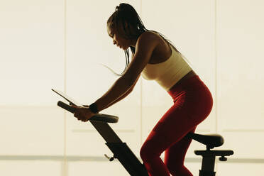 Fit woman, dressed in sports clothing, pedals on an exercise bike in her gym workout routine. Young female demonstrating her commitment to maintaining a healthy lifestyle through regular exercise. - JLPSF30519
