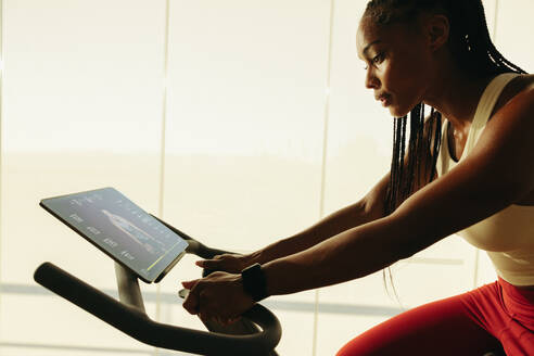 African American woman using a fitness app to guide her exercise bike workout at home. Young black woman taking advantage of smart fitness equipment to achieve a healthy lifestyle. - JLPSF30521